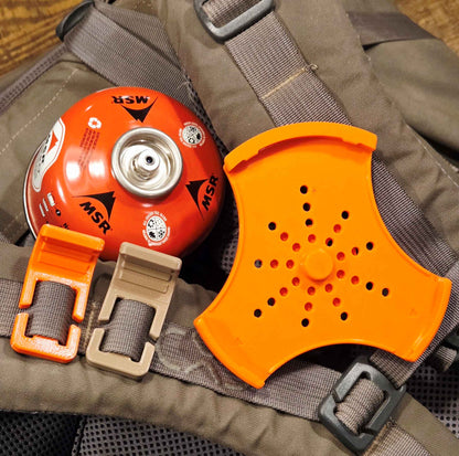 An orange MSR backpacking stove accessory set displayed on a Exo Mountain Gear backpack, including a main stove adapter plate, orange Sling Stop, and a beige Sling Stop, with an orange MSR canister partially visible.